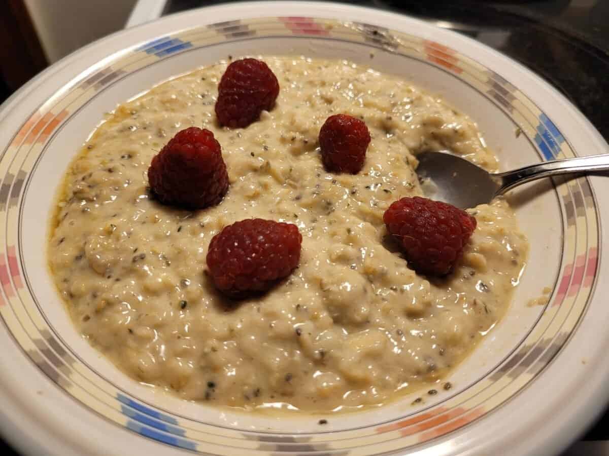 A bowl of oatmeal topped with five raspberries. The oatmeal has visible grains and seeds, indicating a textured mix. A spoon is on the right side of the bowl, which has a decorative rim with red and blue accents.