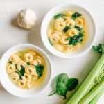Two white bowls of Spinach Tortellini Soup sit on a light surface, surrounded by a head of garlic, fresh spinach leaves, and celery stalks.