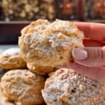 A hand holds a powdered sugar-dusted Apple Cookie (Scone) above a plate of similar treats, with a warm, blurred fireplace in the background, creating a cozy atmosphere.
