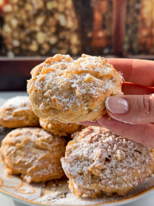 A hand holds a powdered sugar-dusted Apple Cookie (Scone) above a plate of similar treats, with a warm, blurred fireplace in the background, creating a cozy atmosphere.