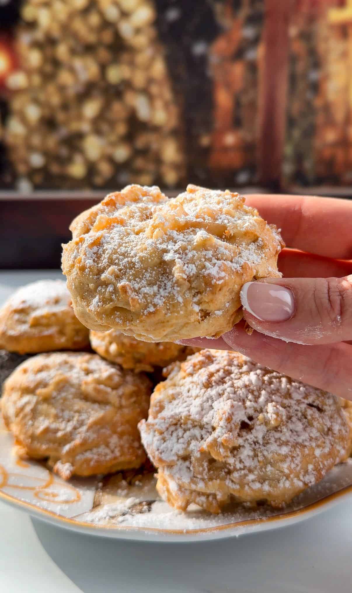 A hand holds a powdered sugar-dusted Apple Cookie (Scone) above a plate of similar treats, with a warm, blurred fireplace in the background, creating a cozy atmosphere.