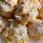 A close-up of several golden, irregularly shaped apple fritters dusted with powdered sugar, sitting on a white surface in bright natural sunlight, inspired by a classic Apple Cookies Recipe.