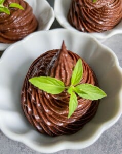A white bowl filled with swirled chocolate mousse, topped with a small sprig of fresh green mint. Hints of crushed Peanut Butter Cups add texture, while two other similar bowls are partially visible in the background.