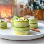 A white plate with neatly stacked Cucumber and Avocado Rolls garnished with sesame seeds, set on a light wooden table. In the blurred background, a decorated Christmas tree and cozy fireplace with stockings and candles add festive warmth.
