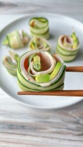 Chopsticks hold a Cucumber and Avocado Roll made with ham, while more cucumber and avocado rolls are arranged on a white plate in the background.