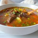 A bowl of beef and vegetable soup with chunks of meat, peas, carrots, and potatoes in a rich broth, garnished with herbs. This hearty Beef Soup Recipe is served with a wooden spoon resting inside.