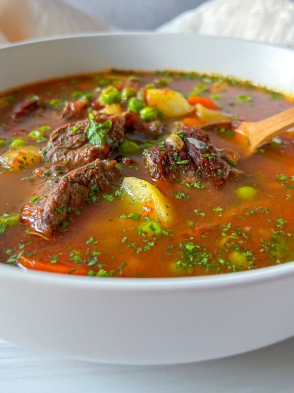 A bowl of beef and vegetable soup with chunks of meat, peas, carrots, and potatoes in a rich broth, garnished with herbs. This hearty Beef Soup Recipe is served with a wooden spoon resting inside.