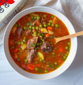A bowl of hearty beef stew with chunks of beef, potatoes, carrots, peas, and fresh herbs sits on a white surface—perfect comfort food, much like a cozy Spinach Tortellini Soup. Served with a wooden spoon for rustic charm.