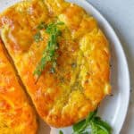 Golden, freshly baked bread topped with herbs and a sprig of fresh thyme, reminiscent of Bulgarian Cheese Sandwiches (Prinzesi), served on a white plate; part of another bread loaf and green herbs are visible on the side.
