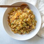 A white bowl filled with a Deconstructed Cabbage Rolls mixture of cooked cabbage and ground meat, garnished with chopped herbs, sits on a light surface with a wooden spoon resting on the rim. A beige cloth is partially visible beside the bowl.