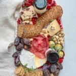 A Holiday Charcuterie Board with assorted cheeses, crackers, salami, prosciutto, grapes, dates, pistachios, pomegranate seeds, two jars of spreads, and fresh herbs is beautifully arranged on a white surface with a napkin.