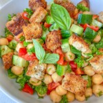 A bowl of Chickpea Crouton Salad with diced cucumbers, tomatoes, fresh parsley, crunchy croutons, and basil leaves, all beautifully arranged in a white bowl.
