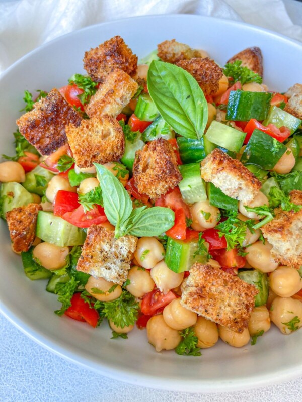 A bowl of Chickpea Crouton Salad with diced cucumbers, tomatoes, fresh parsley, crunchy croutons, and basil leaves, all beautifully arranged in a white bowl.