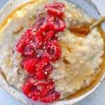 A bowl of creamy custard oatmeal topped with fresh raspberries, mixed seeds, and a drizzle of honey or syrup. A gold spoon rests in the bowl, which sits on a light wooden surface.