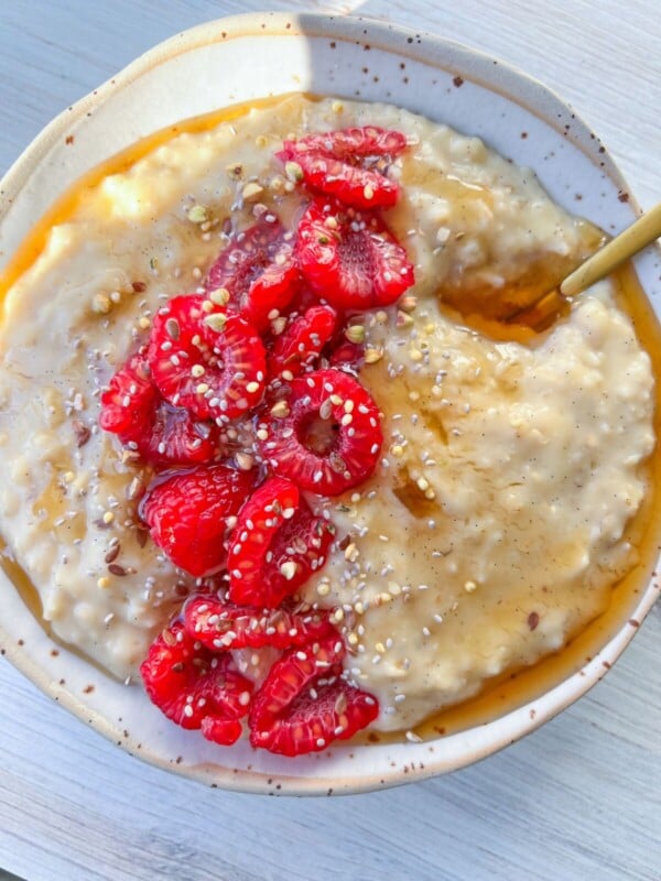 A bowl of creamy custard oatmeal topped with fresh raspberries, mixed seeds, and a drizzle of honey or syrup. A gold spoon rests in the bowl, which sits on a light wooden surface.