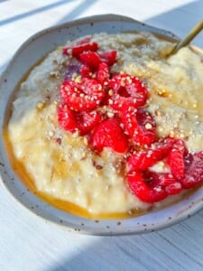 A bowl of creamy custard oatmeal topped with fresh raspberries, seeds, and a drizzle of honey, served in a shallow ceramic bowl on a light-colored table in bright natural light.