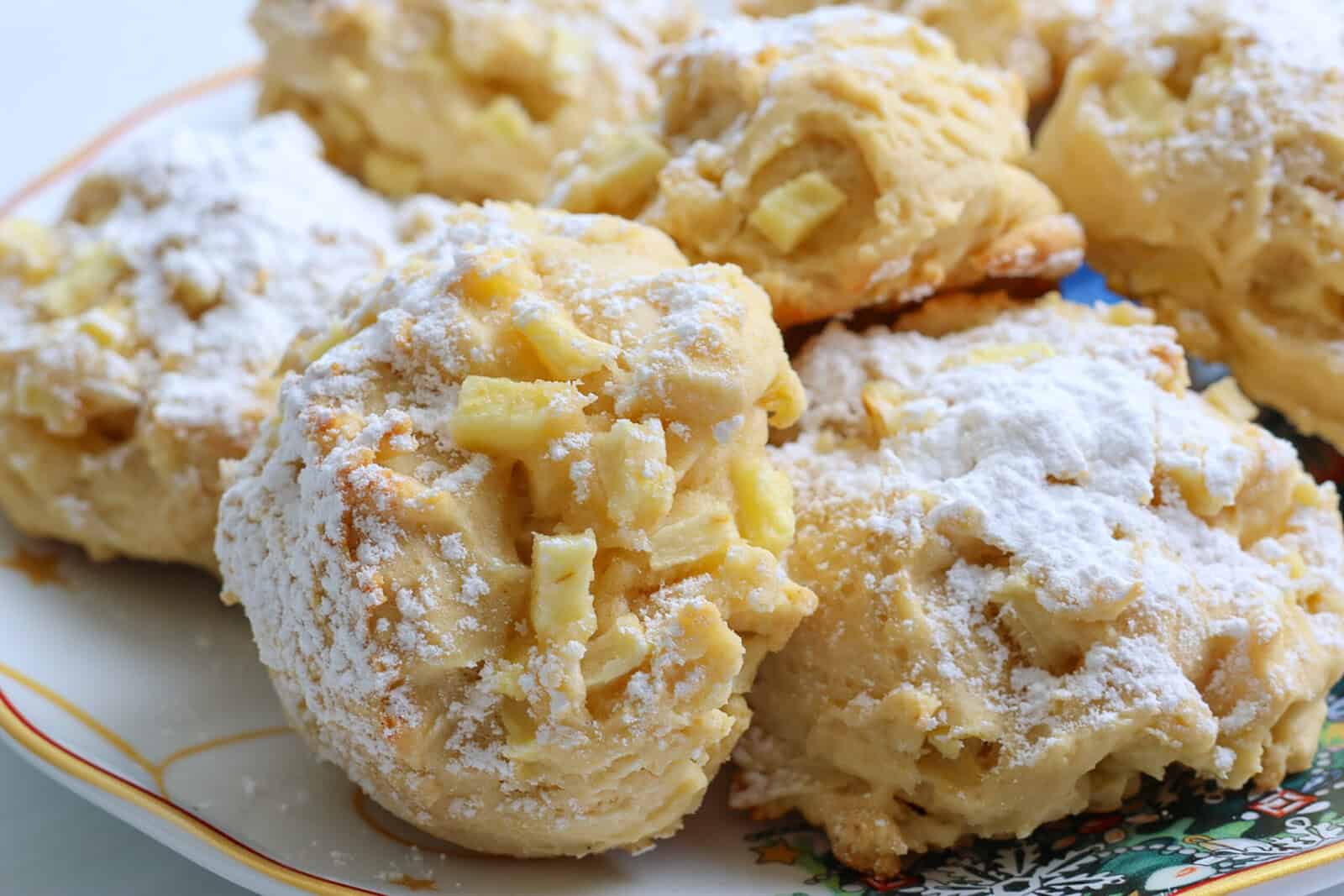 Close-up of several golden Apple Cookies (Scones) sprinkled with powdered sugar, arranged on a decorative plate with a colorful pattern in the corner. The cookies have a chunky texture and visible pieces of apple.