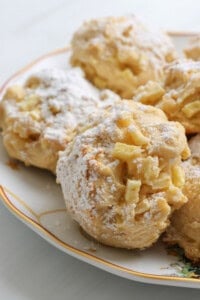 Close-up of apple fritters and Apple Cookies (Scones) dusted with powdered sugar, arranged on a decorative plate. The fritters have a golden, crispy texture with visible apple pieces.
