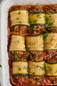 Close-up of baked zucchini rolls stuffed with a savory mixture, arranged in a white casserole dish. The rolls are topped with chopped parsley and set on a tomato-based sauce, with some browned edges visible on the dish.