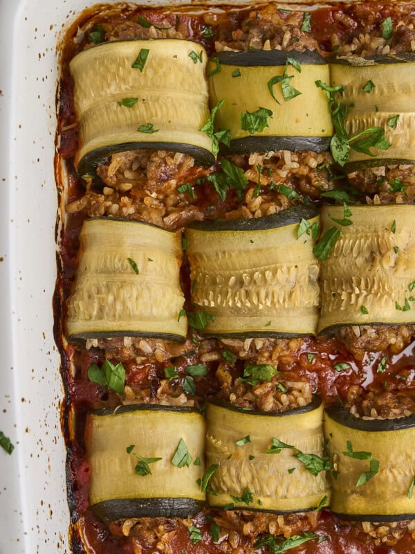 Close-up of baked zucchini rolls stuffed with a savory mixture, arranged in a white casserole dish. The rolls are topped with chopped parsley and set on a tomato-based sauce, with some browned edges visible on the dish.