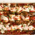 A baked dish of rolled zucchini slices filled with a savory mixture, topped with tomato sauce, melted cheese, and fresh herbs, all in a white rectangular baking dish. The dish appears freshly cooked and garnished.