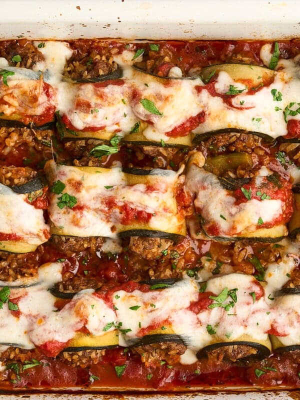 A baked dish of rolled zucchini slices filled with a savory mixture, topped with tomato sauce, melted cheese, and fresh herbs, all in a white rectangular baking dish. The dish appears freshly cooked and garnished.