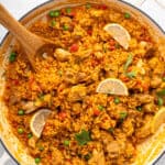 A large pan with flavorful chicken and rice dish, featuring golden rice mixed with pieces of chicken, green peas, and red bell peppers. Topped with parsley and lemon slices, a wooden spoon rests on the side.