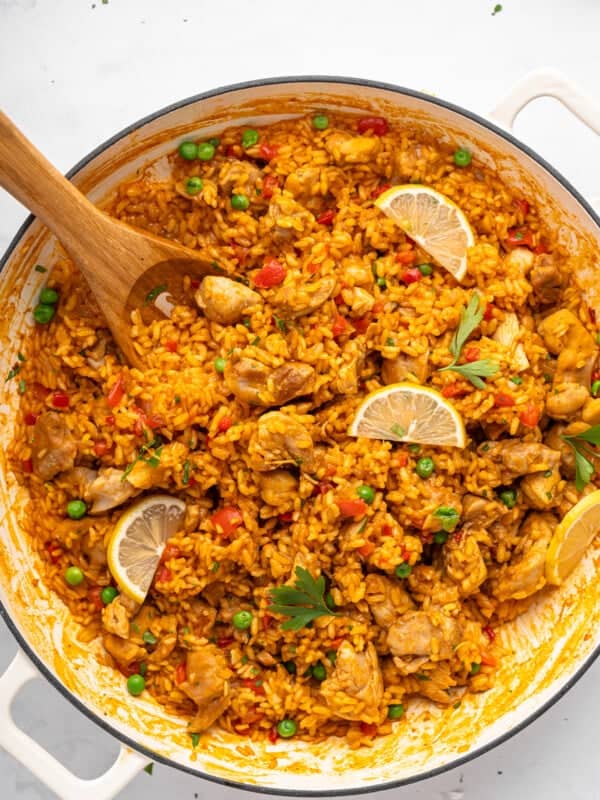A large pan with flavorful chicken and rice dish, featuring golden rice mixed with pieces of chicken, green peas, and red bell peppers. Topped with parsley and lemon slices, a wooden spoon rests on the side.