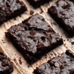 Close-up of several rich, chocolate brownies topped with chocolate chips, arranged on parchment paper. The brownies have a dense, fudgy texture, and the chocolate chips add a touch of shine.