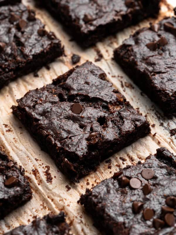 Close-up of several rich, chocolate brownies topped with chocolate chips, arranged on parchment paper. The brownies have a dense, fudgy texture, and the chocolate chips add a touch of shine.