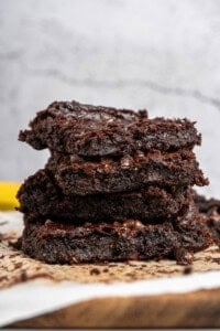 A close-up of a stack of three gooey chocolate brownies on parchment paper. The brownies are rich and moist, with visible chocolate chips embedded in the batter. The background is a simple, textured surface.