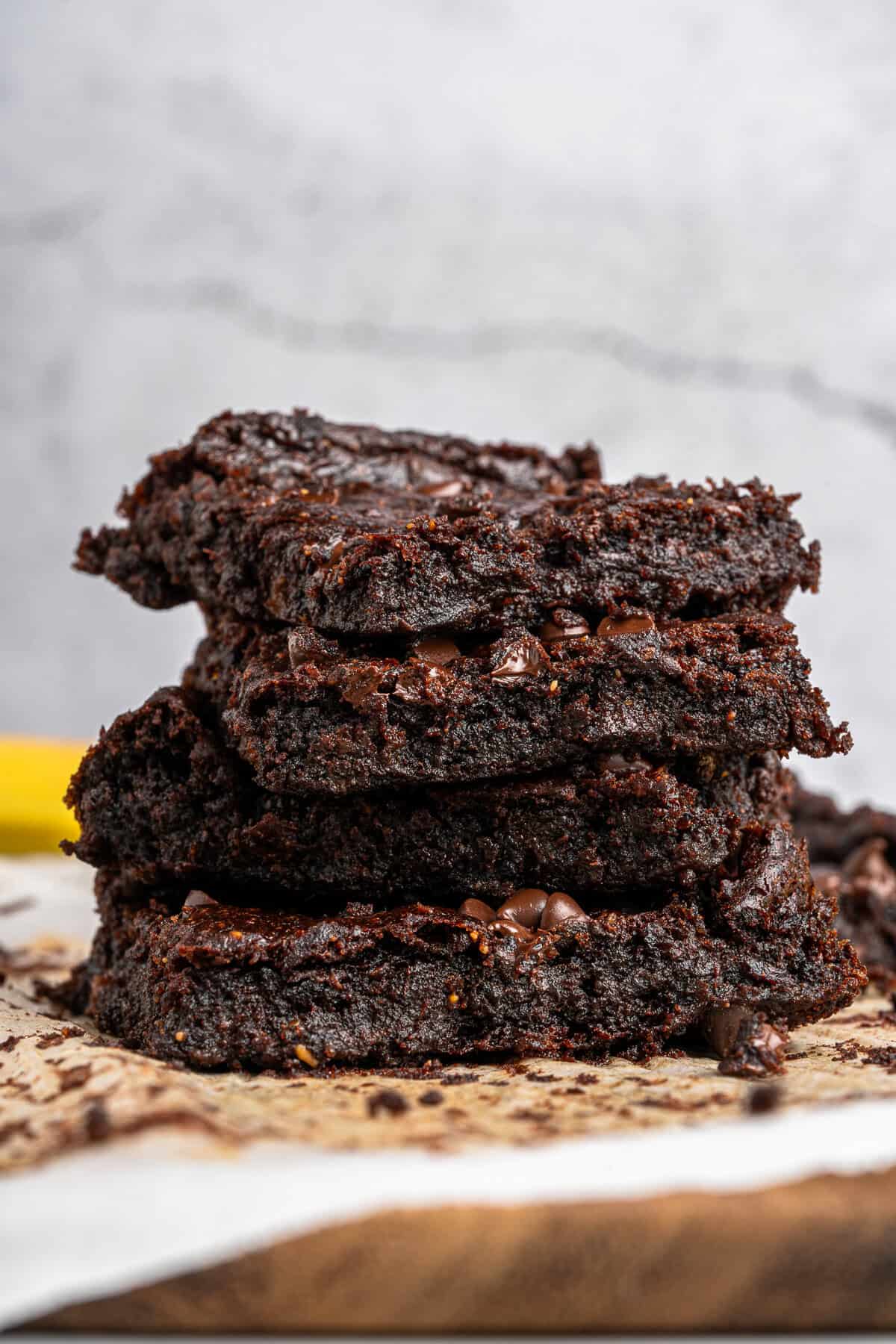 A close-up of a stack of three gooey chocolate brownies on parchment paper. The brownies are rich and moist, with visible chocolate chips embedded in the batter. The background is a simple, textured surface.