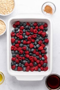 A white baking dish filled with fresh raspberries and blueberries. Surrounding the dish are bowls containing oats, flour, cinnamon, olive oil, and syrup. The ingredients are arranged on a light-colored surface.