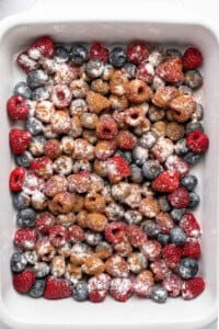A white baking dish filled with fresh raspberries and blueberries, sprinkled with powdered sugar and cinnamon.