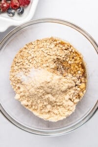 A glass bowl filled with oats, almond flour, and brown sugar, ready for mixing. On the side, a dish with assorted berries adds a colorful contrast. The ingredients are set against a white background, suggesting preparation for a recipe.