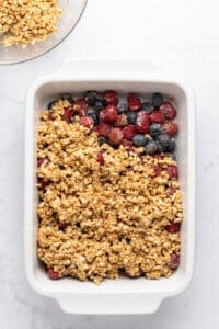 A white baking dish with a partially covered layer of mixed berries, including raspberries and blueberries, topped with uncooked oat crumble mixture. There is a glass bowl with more oats in the top left corner.