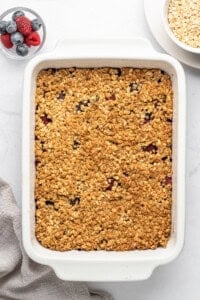A baked oatmeal dish in a white rectangular casserole dish on a marble surface. Next to it are a bowl of mixed berries and a bowl of oats. A folded beige cloth is placed nearby.