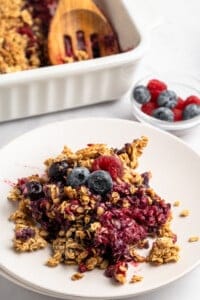 A plate with a serving of berry crisp topped with granola and fresh blueberries and raspberries. In the background, a baking dish with more crisp and a small bowl of berries are visible.