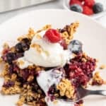 A serving of mixed berry crisp topped with a scoop of vanilla ice cream and garnished with a raspberry on a white plate. The crisp contains oats and various berries, with a fork resting on the plate. A dish of additional berries is in the background.