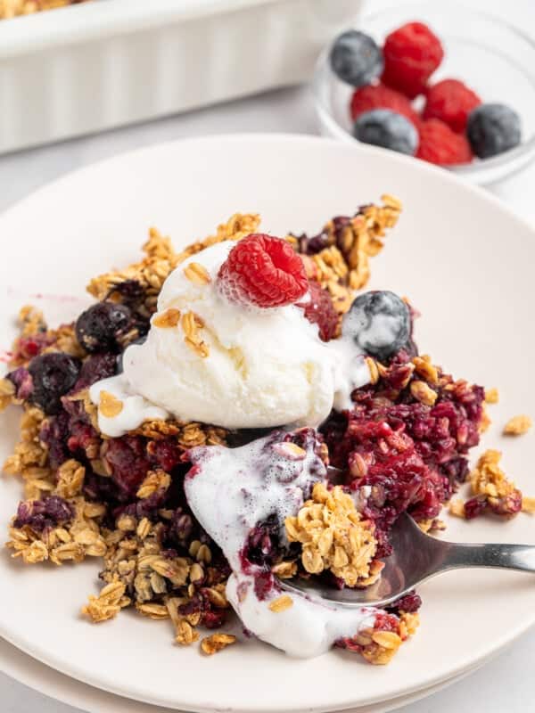 A serving of mixed berry crisp topped with a scoop of vanilla ice cream and garnished with a raspberry on a white plate. The crisp contains oats and various berries, with a fork resting on the plate. A dish of additional berries is in the background.