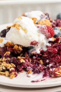 A close-up of a berry crumble topped with a scoop of vanilla ice cream. The crumble contains oatmeal, blueberries, and raspberries, with melted ice cream drizzling over the dessert on a white plate.