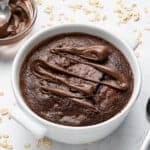 A baked oatmeal dish topped with drizzled chocolate sits in a white bowl. A spoon rests beside it on a white surface scattered with oats. A bowl of chocolate spread is nearby.