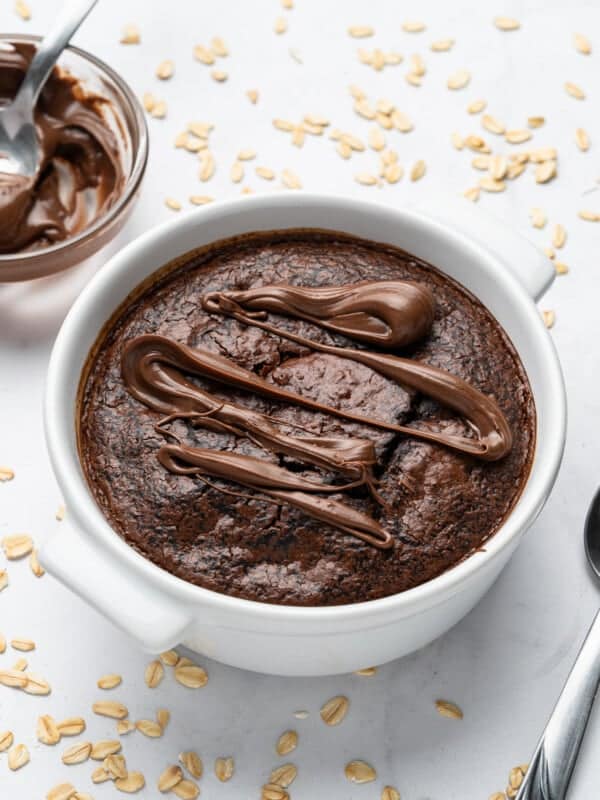 A baked oatmeal dish topped with drizzled chocolate sits in a white bowl. A spoon rests beside it on a white surface scattered with oats. A bowl of chocolate spread is nearby.