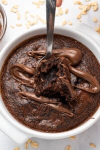 A spoon taking a bite out of a chocolate dessert in a white dish, topped with swirls of melted chocolate. Oats are scattered around the dish on a light surface.