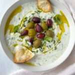 A bowl of creamy tzatziki made from an easy tzatziki recipe, topped with green and purple olives, fresh herbs, a drizzle of olive oil, and pita chips for garnish, all placed on a light cloth.