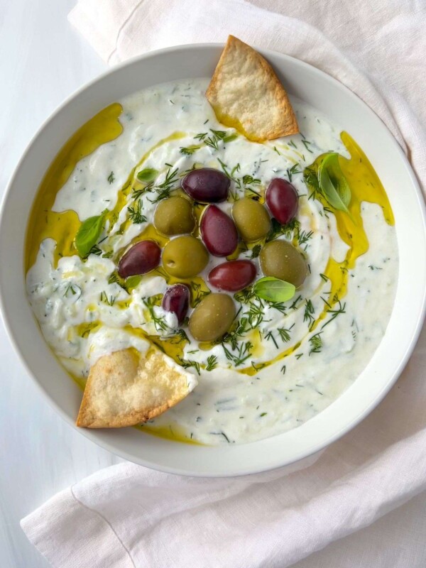 A bowl of creamy tzatziki made from an easy tzatziki recipe, topped with green and purple olives, fresh herbs, a drizzle of olive oil, and pita chips for garnish, all placed on a light cloth.