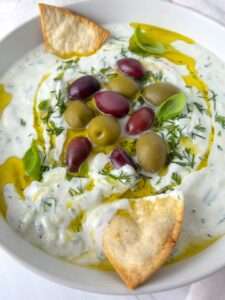 A bowl of creamy Easy Tzatziki Recipe sauce topped with mixed olives, fresh dill, basil leaves, a drizzle of olive oil, and two triangular pita chips around the edge.