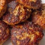 A close-up of several golden-brown, pan-fried sausage patties stacked on a white plate. The crispy, caramelized exterior and juicy texture make them perfect for anyone looking for an Easy Meatball Recipe alternative.