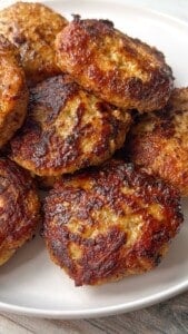A close-up of several golden-brown, pan-fried sausage patties stacked on a white plate. The crispy, caramelized exterior and juicy texture make them perfect for anyone looking for an Easy Meatball Recipe alternative.