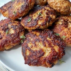 A white plate filled with several browned, grilled chicken patties—resembling an easy meatball recipe—garnished with small sprigs of fresh herbs, set on a light-colored wooden surface.